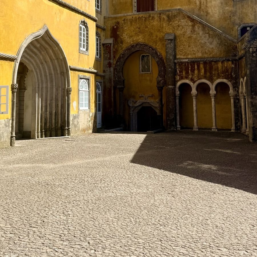 Sunlit courtyard with a Gothic arch, a Moorish arch, and a hard diagonal shadow falling across the cobblestones, Pena Palace, Sintra, Portugal