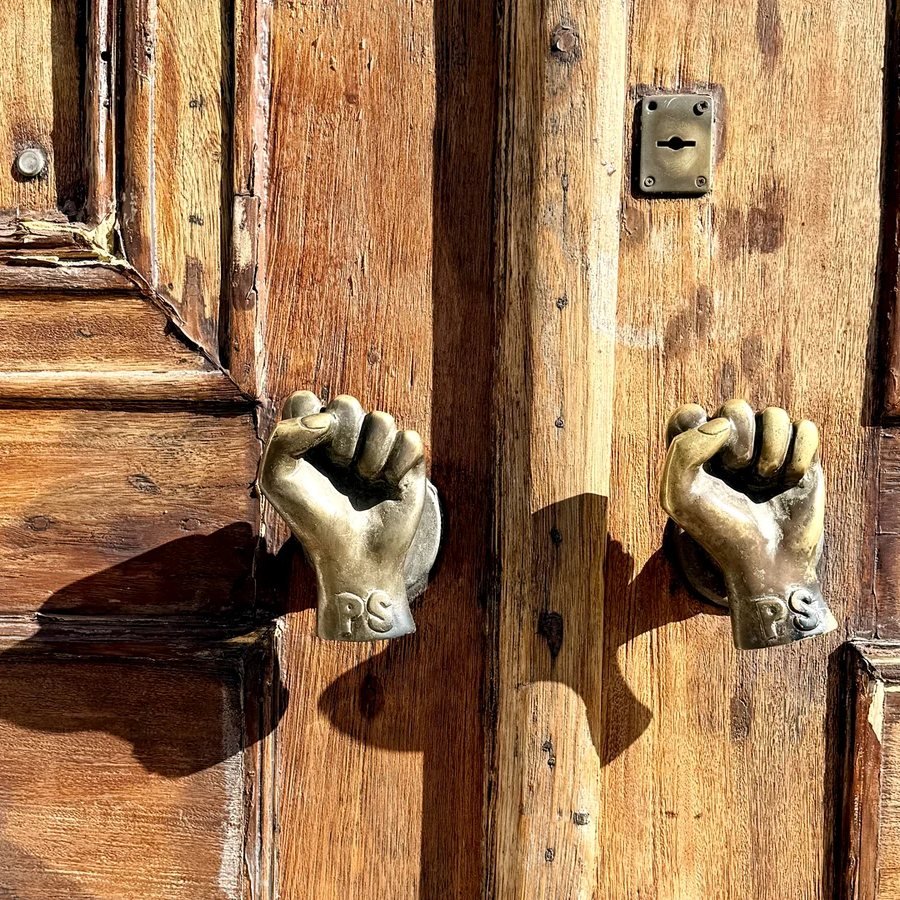 Weathered wooden double door in Lisbon with two cast brass fist knockers throwing hard afternoon shadows - 3½ Things September 2026 Edition cover